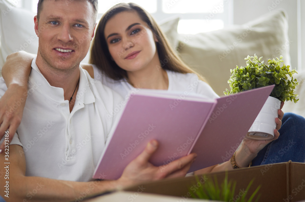 Cute couple unpacking cardboard boxes in their new home, sitting on the floor and looking at a family album Cute couple unpacking cardboard boxes in their new home, sitting on the floor and looking at a family album