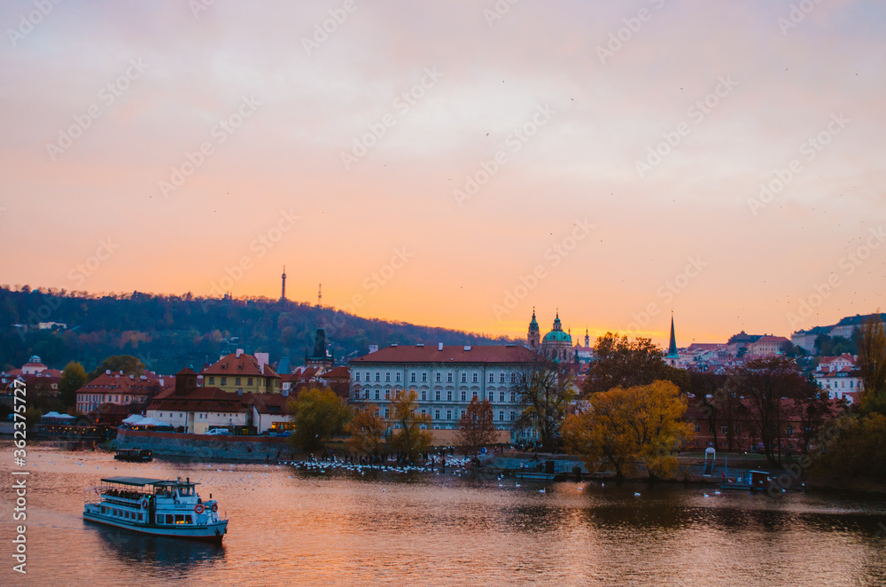 Naklejka premium Dramatic sundown in the Vltava River with a boat passing by and a gradient of orange to pink colors reflecting from the sky into the water.