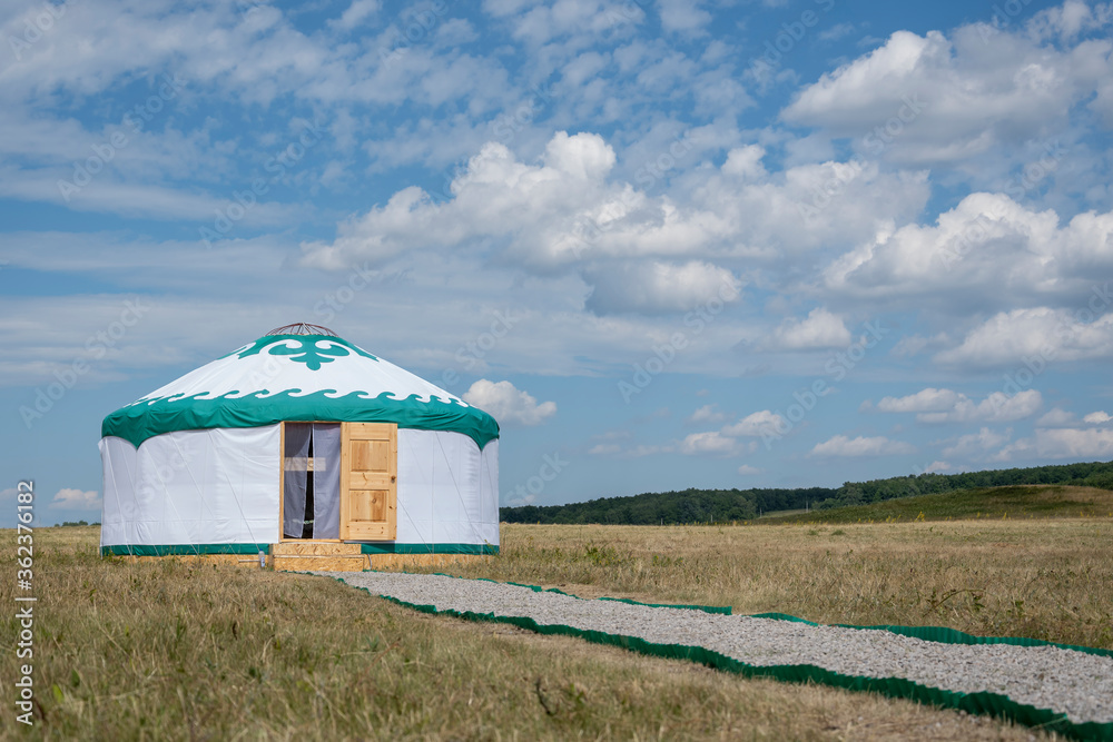 A white yurt (the house of nomadic people, Bashkiria) stands in the steppe with an open door against a blue summer sky