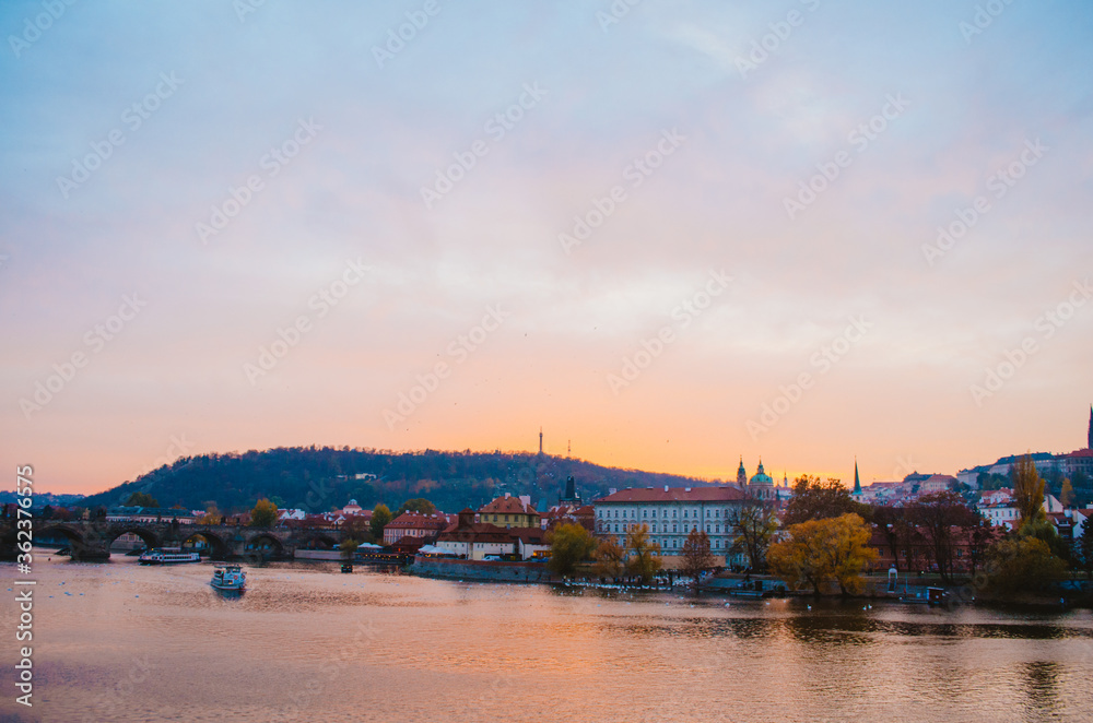Dramatic sundown in the Vltava River with a boat passing by and a gradient of orange to pink colors reflecting from the sky into the water.