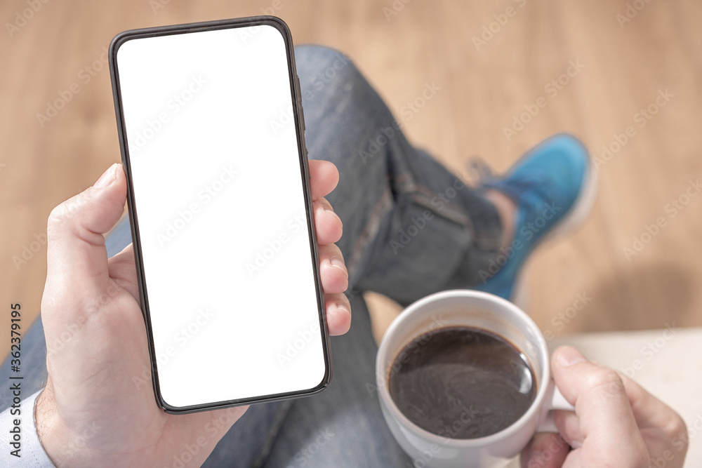 Top view mockup image of a man holding a black mobile phone with blank white desktop screen with coffee cup