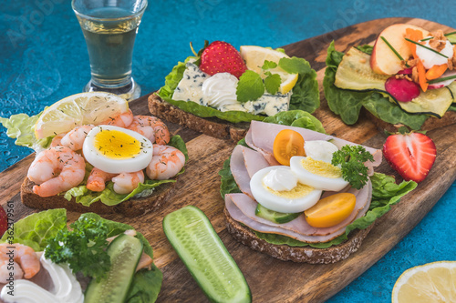 Variety of danish open sandwiches on wooden boards and blue background, decorated with vegetables, strawberries and aquavit