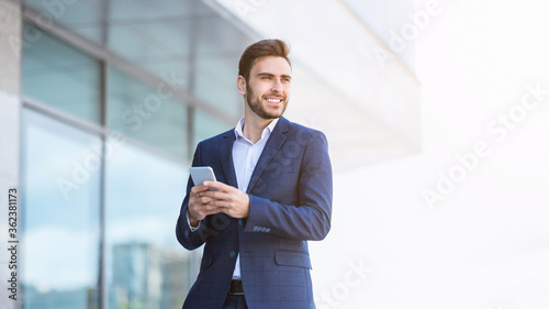 Fotografie Smiling millennial industrialist checking his smartphone near skyscraper on city