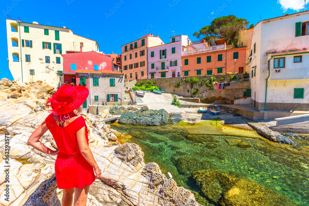 Woman in red dress over cliffs at Marciana Marina looking famous houses of old village Borgo al Cotone in bay of Marciana Marina seaside. Elegant tourist on holiday travel on Elba Island, Italy.