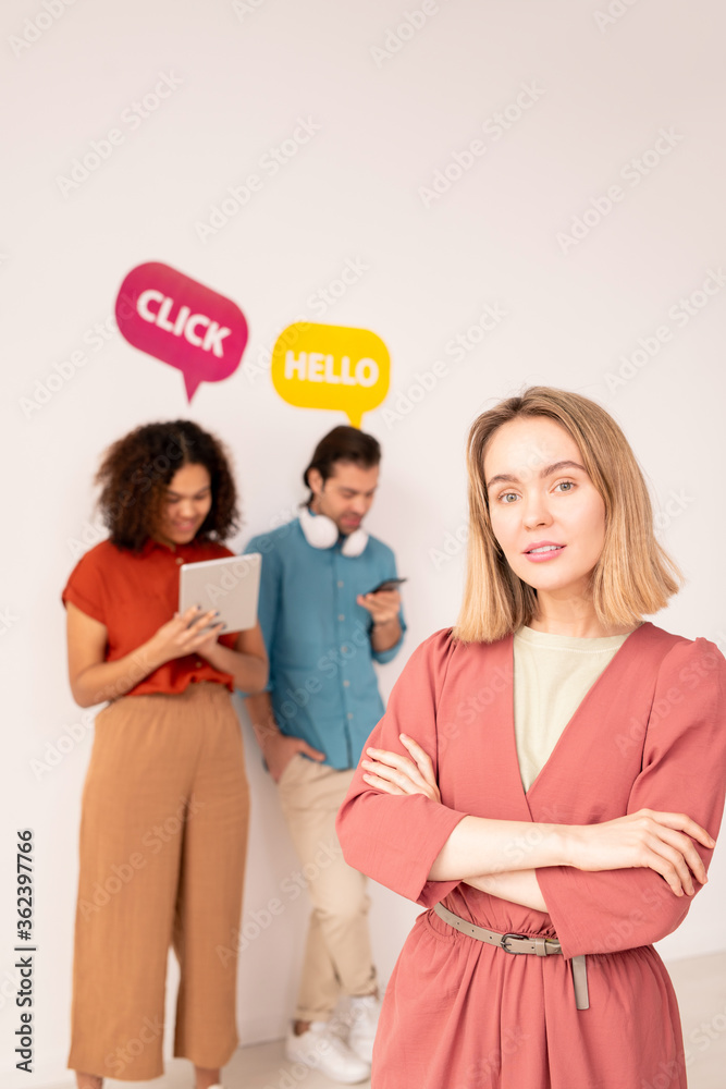 Young blond cross-armed female stanging against friends using mobile gadgets Young blond cross-armed female stanging against friends using mobile gadgets