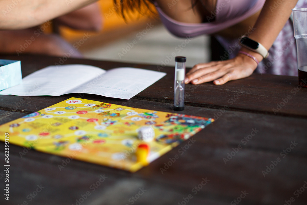Board game board, chips, cube, timer on a dark wooden table and hands in the background. The concept of teamwork, intellectual relaxation, corporate event, playing at home with children. Copy space Board game board, chips, cube, timer on a dark wooden table and hands in the background. The concept of teamwork, intellectual relaxation, corporate event, playing at home with children. Copy space