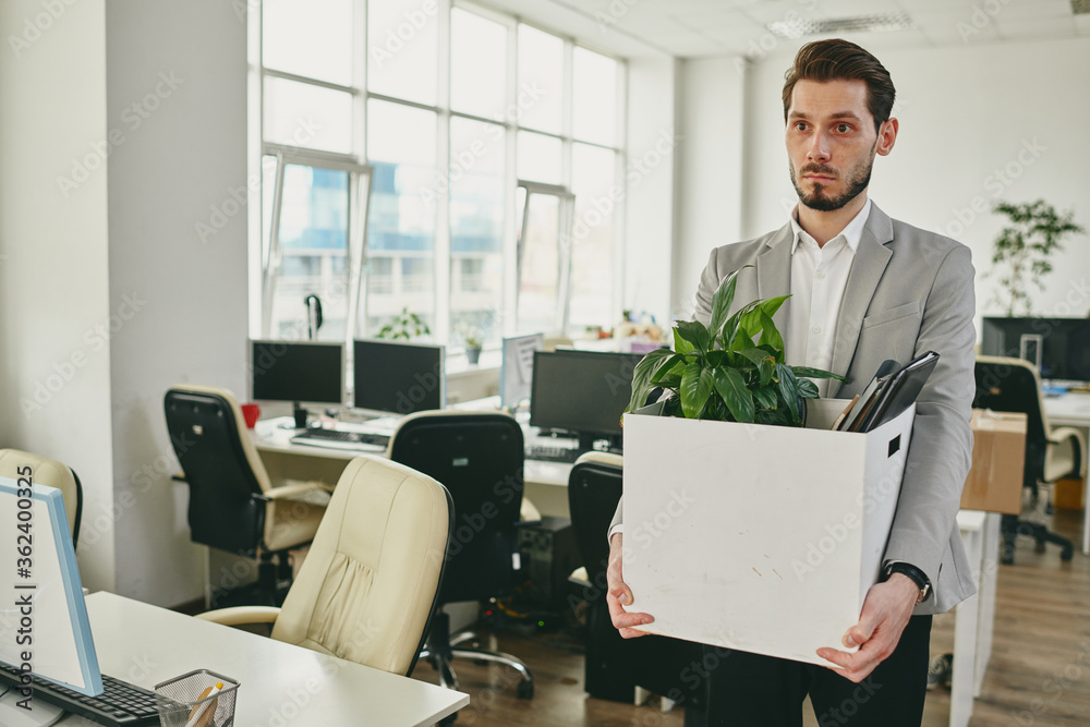 Sad young man carrying box with green plant and documents while leaving office Sad young man carrying box with green plant and documents while leaving office