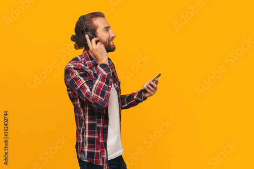 Young  happy man listens to music on headphones  holds a smartphone in his hands  looks away and smiles  on a yellow background