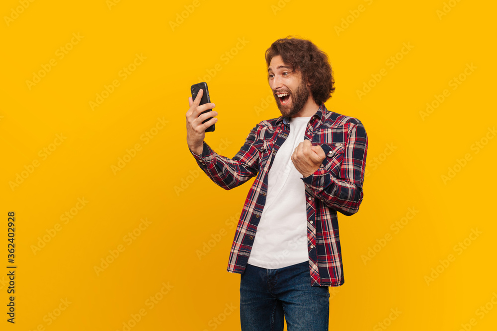 Happy young man rejoices in victory, looking at the phone on a yellow background. A man with long hair and a shirt won the lottery Happy young man rejoices in victory, looking at the phone on a yellow background. A man with long hair and a shirt won the lottery