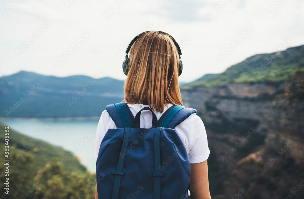 view from behind girl backpacker with blonde hair listening to music headphones standing peak in rocky mountains, traveler with backpack looks into distance enjoying view river valley, copy space