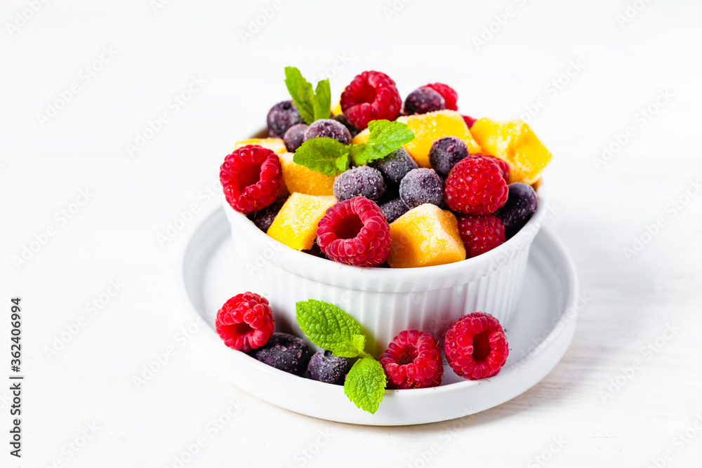 Frozen fruits: mango, raspberry, blueberry with fresh green mint in white bowl. Concept of healthy eating, delicious low calories dessert, summer refreshing light meal. White background copy space Frozen fruits: mango, raspberry, blueberry with fresh green mint in white bowl. Concept of healthy eating, delicious low calories dessert, summer refreshing light meal. White background copy space