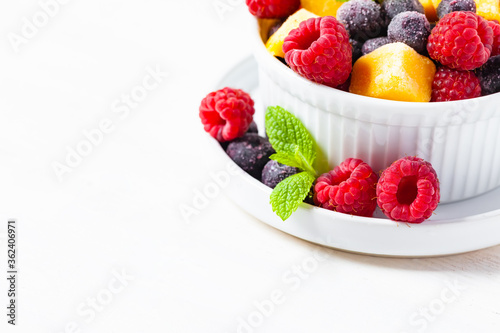 Frozen fruits  mango  raspberry  blueberry with fresh green mint in white bowl. Concept of healthy eating  delicious low calories dessert  summer refreshing light meal. White background copy space