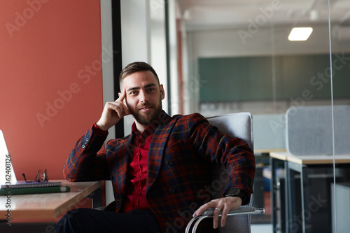 Portrait of elegant bearded businessman looking at camera and smiling while sitting at desk and posing against red wall in office, copy space