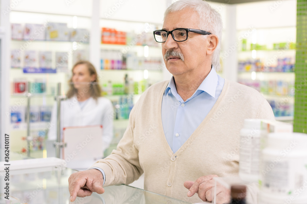Senior client of drugstore standing by counter and waiting for pharmacist Senior client of drugstore standing by counter and waiting for pharmacist