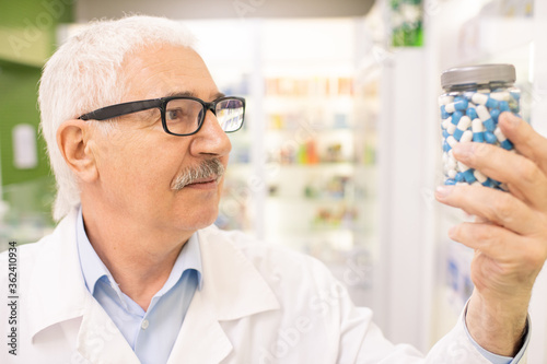 Aged male pharmacist or consultant of large drugstore looking at jar with pills