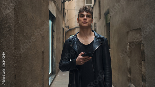 Young attractive man with smartphone thoughtfully looking away on narrow street photo