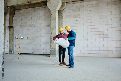 Two young engineers or builders in hardhats and casualwear looking at blueprint