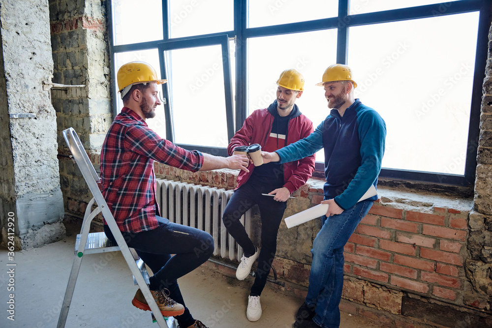 Three young architects in protective helmets clinking with glasses of coffee