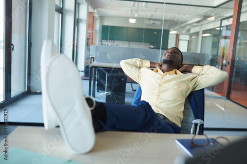 Portrait of contemporary African-American businessman relaxing at workplace in office with feet on desk  copy space