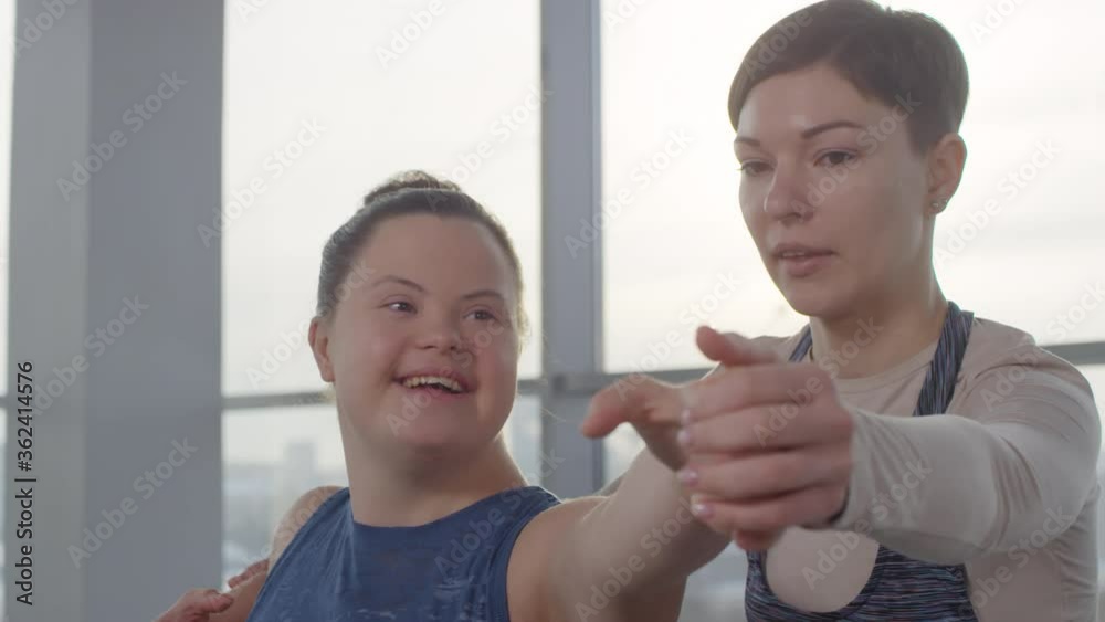 Cheerful young disabled woman with down syndrome doing yoga exercise with her personal trainer helping her Cheerful young disabled woman with down syndrome doing yoga exercise with her personal trainer helping her