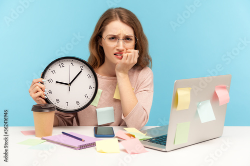 Upset nervous woman employee sitting at workplace office, all covered with sticky notes and holding big clock, looking with anxiety, biting nails. indoor studio shot isolated on blue background
