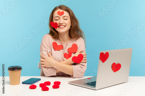 Charming happy young woman with kind smile sitting at workplace office, all covered with sticker love hearts, embracing herself, enjoying valentine's day greetings. indoor studio shot, isolated