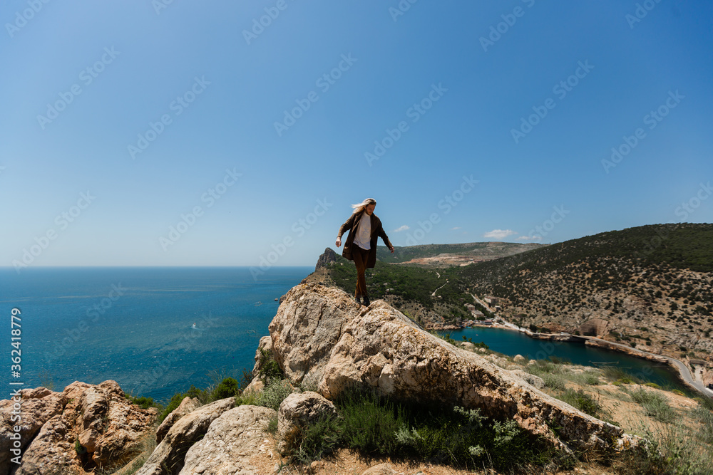 A man in brown clothes with blond hair blowing in the wind, walks along the edge of a semicircular rock helping himself with his hands to balance against the background of the blue sea under the sun