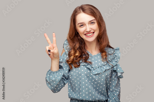 Optimistic happy joyful girl ruffle blouse showing victory peace gesture two raised fingers, looking at camera with toothy smile, enjoying win victory. indoor studio shot isolated on gray background