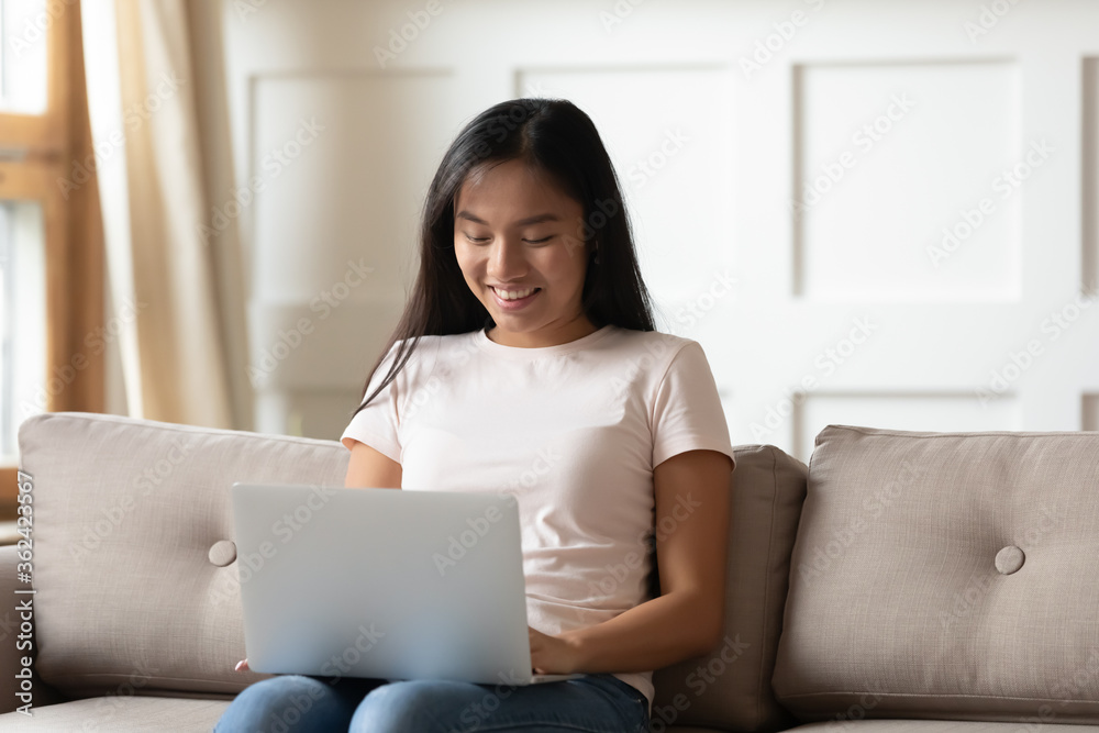 Smiling young vietnamese pretty woman sitting on sofa, holding computer on lap, enjoying web surfing internet, using laptop applications, communicating with friends online, shopping in internet store. Smiling young vietnamese pretty woman sitting on sofa, holding computer on lap, enjoying web surfing internet, using laptop applications, communicating with friends online, shopping in internet store.