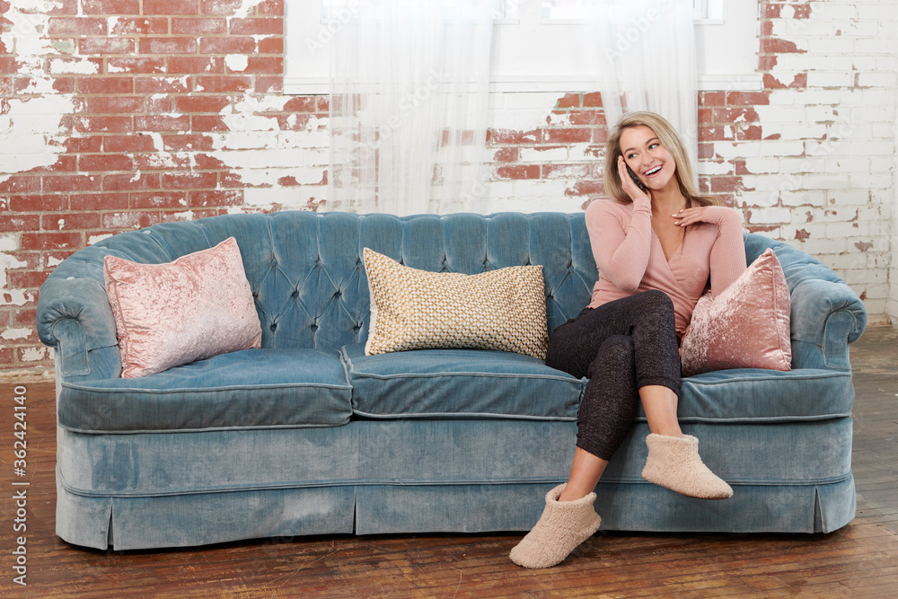 Obraz premium Stunning young blonde woman in pink blouse and dark comfortable pants relaxes on couch and talks on cell phone