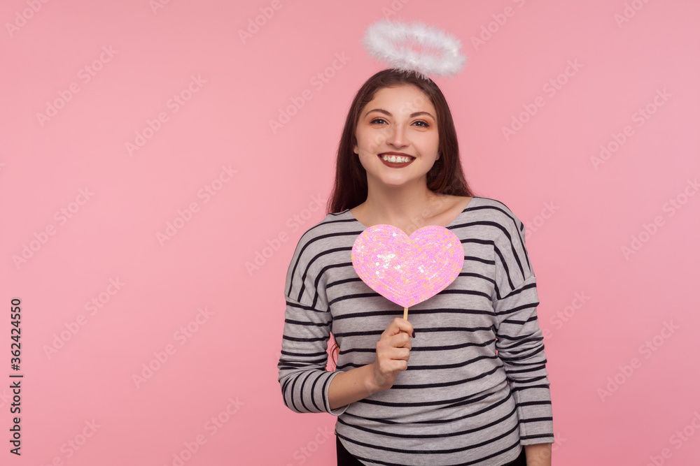 Happy Valentine's day! Angelic woman with holy nimbus holding pink paper heart and smiling to camera, demonstrating love care, romantic feelings. indoor studio shot isolated on pink background Happy Valentine's day! Angelic woman with holy nimbus holding pink paper heart and smiling to camera, demonstrating love care, romantic feelings. indoor studio shot isolated on pink background