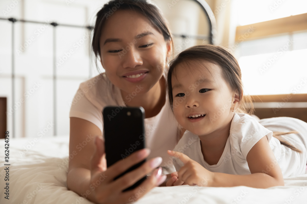 Head shot close up happy young vietnamese woman lying on bed with cute small biracial kid daughter, posing for selfie photo on smartphone or recording video for social network blog in bedroom. Head shot close up happy young vietnamese woman lying on bed with cute small biracial kid daughter, posing for selfie photo on smartphone or recording video for social network blog in bedroom.