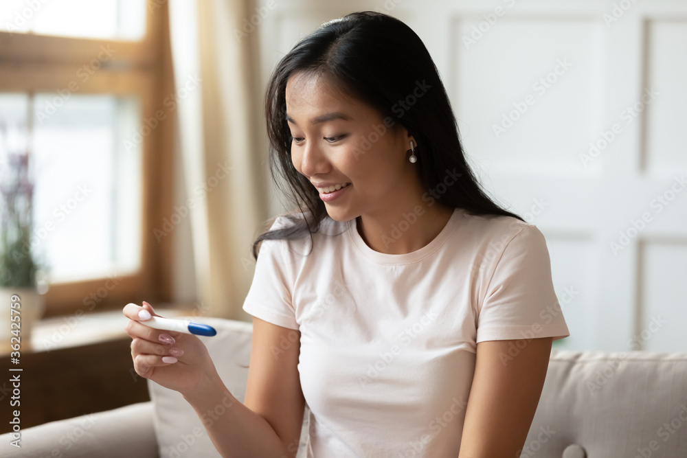 Smiling young excited attractive vietnamese ethnicity woman sitting on sofa, looking at plastic pregnancy test, waiting for results. happy asian lady checking ovulation, future maternity concept. Smiling young excited attractive vietnamese ethnicity woman sitting on sofa, looking at plastic pregnancy test, waiting for results. happy asian lady checking ovulation, future maternity concept.