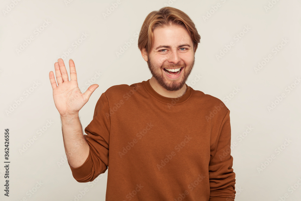 Portrait of hospitable friendly bearded man in sweatshirt raising hand waving hello hi and smiling, happy to meet friend, welcoming or gesturing goodbye. indoor studio shot isolated on gray background Portrait of hospitable friendly bearded man in sweatshirt raising hand waving hello hi and smiling, happy to meet friend, welcoming or gesturing goodbye. indoor studio shot isolated on gray background
