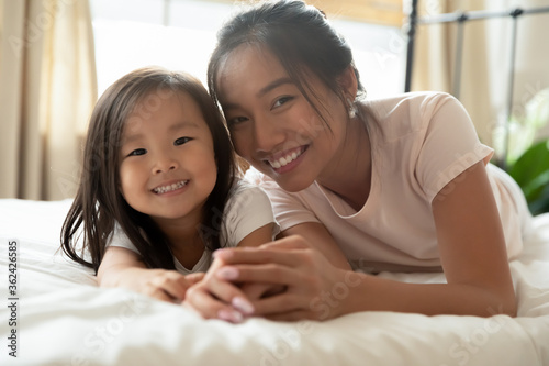 Head shot close up portrait adorable small vietnamese baby girl lying on comfortable bed with smiling young mommy, happy asian different generations family relaxing in bedroom, looking at camera.