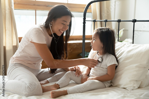 Caring affectionate smiling vietnamese ethnicity mother using stethoscope, listening heartbeat or lungs of little preschool baby girl in bedroom, curing illness or playing doctor patient on bed.