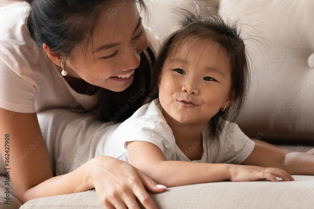 Happy young caring vietnamese mommy cuddling adorable funny small biracial baby girl, lying together on comfortable couch, head shot different generations cheerful family enjoying sweet tender moment. Happy young caring vietnamese mommy cuddling adorable funny small biracial baby girl, lying together on comfortable couch, head shot different generations cheerful family enjoying sweet tender moment.