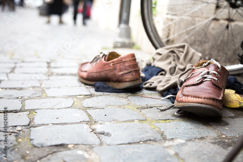 Urban street composition. Pair of brown leather boots with white shoelaces, light beige pullover on cobblestone pavement. loafers thrown in the city. Bicycle wheel in the background. Fashion Garbage 