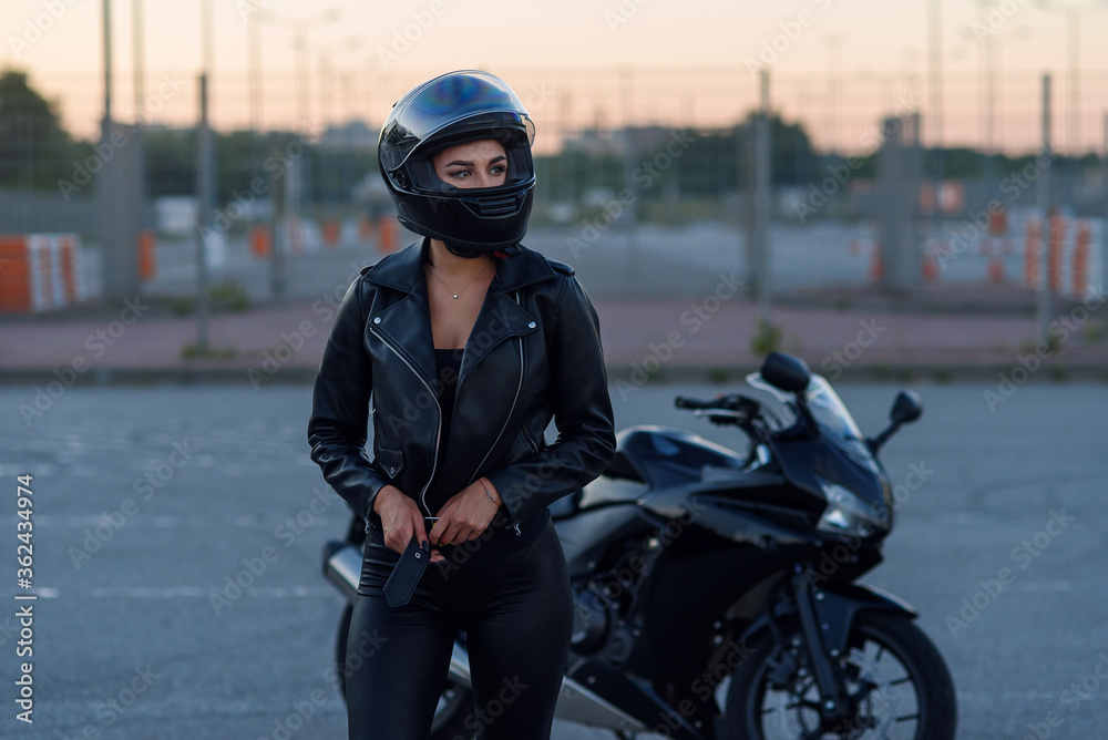 Biker woman in black leather jacket and full face helmet stands near stylish sports motorcycle. Urban parking, sunset in big city. Traveling and active hipster lifestyle. Girls power. Biker woman in black leather jacket and full face helmet stands near stylish sports motorcycle. Urban parking, sunset in big city. Traveling and active hipster lifestyle. Girls power.