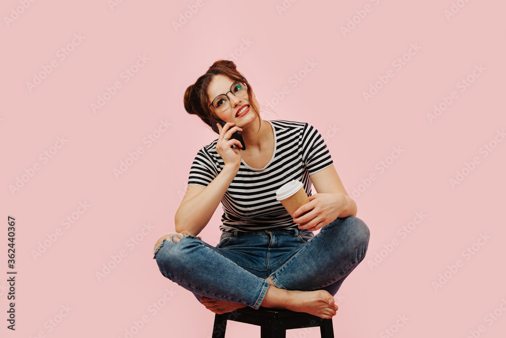 Young woman sitting on a stool, keen on talking on phone with coffee in hand Young woman sitting on a stool, keen on talking on phone with coffee in hand
