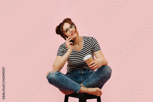 Young woman sitting on a stool, keen on talking on phone with coffee in hand