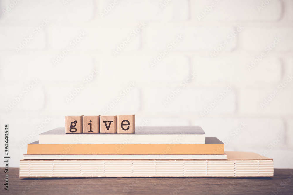 The word GIVE, letters on block cubes on top of books and wooden table with white bricks background, blank copy space, vintage minimal style. Concepts of share, charity, donation and love. The word GIVE, letters on block cubes on top of books and wooden table with white bricks background, blank copy space, vintage minimal style. Concepts of share, charity, donation and love.