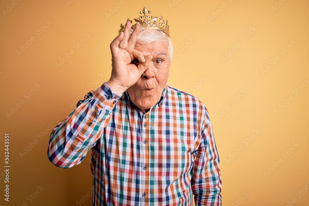 Senior handsome hoary man wearing golden crown of king over isolated yellow background doing ok gesture shocked with surprised face, eye looking through fingers. Unbelieving expression. Senior handsome hoary man wearing golden crown of king over isolated yellow background doing ok gesture shocked with surprised face, eye looking through fingers. Unbelieving expression.