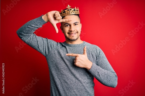 Young handsome man wearing golden crown of prince over isolated red background smiling making frame with hands and fingers with happy face. Creativity and photography concept. photo