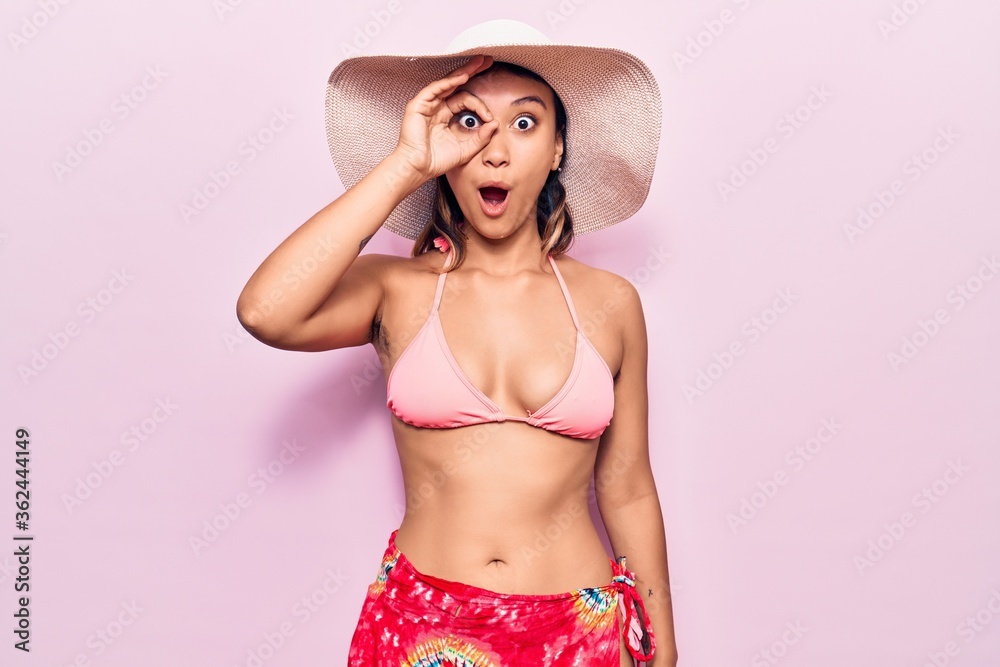 Young woman wearing bikini and hat doing ok gesture shocked with surprised face, eye looking through fingers. unbelieving expression. Young woman wearing bikini and hat doing ok gesture shocked with surprised face, eye looking through fingers. unbelieving expression.