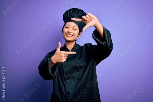 Young beautiful chinese chef woman wearing cooker uniform and hat over purple background smiling making frame with hands and fingers with happy face. Creativity and photography concept.