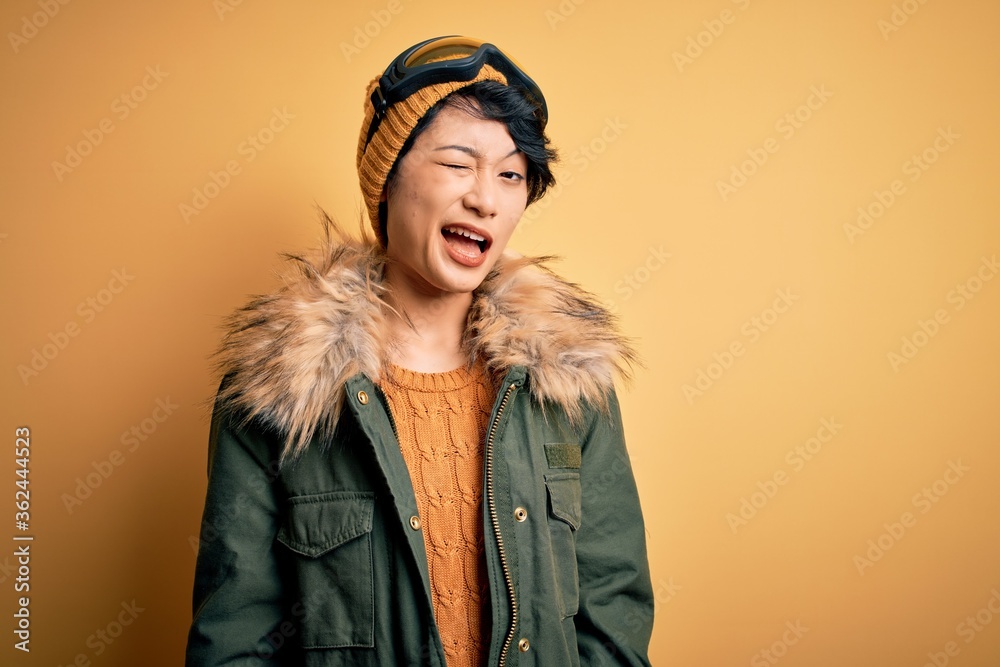Beautiful asian skier girl wearing snow sportswear using ski goggles over yellow background winking looking at the camera with sexy expression, cheerful and happy face. Beautiful asian skier girl wearing snow sportswear using ski goggles over yellow background winking looking at the camera with sexy expression, cheerful and happy face.