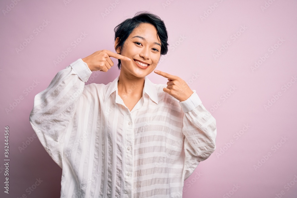Young beautiful asian girl wearing casual shirt standing over isolated pink background smiling cheerful showing and pointing with fingers teeth and mouth. Dental health concept. Young beautiful asian girl wearing casual shirt standing over isolated pink background smiling cheerful showing and pointing with fingers teeth and mouth. Dental health concept.
