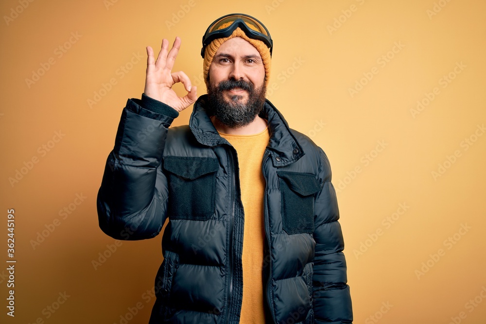 Handsome skier bald man with beard skiing wearing snow sportswear and ski goggles smiling positive doing ok sign with hand and fingers. Successful expression. Handsome skier bald man with beard skiing wearing snow sportswear and ski goggles smiling positive doing ok sign with hand and fingers. Successful expression.