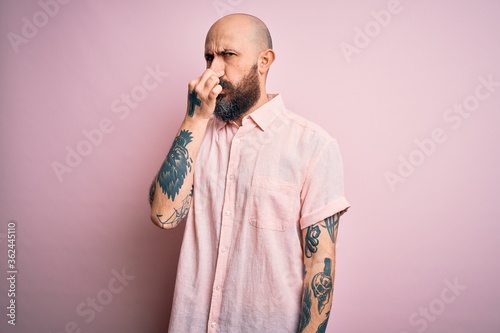Handsome bald man with beard and tattoo wearing casual shirt over isolated pink background smelling something stinky and disgusting, intolerable smell, holding breath with fingers on nose. Bad smell